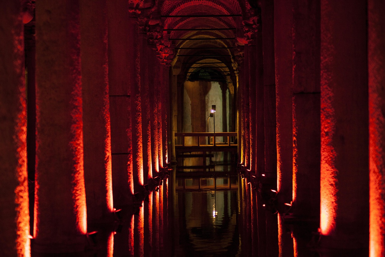 Eine atmosphärische Innenaufnahme der Istanbuler Basilica Cistern (Yerebatan Sarnıcı), die das Gefühl von "Mystik unter der Erde" perfekt einfängt. Reihen von massiven Säulen, die aus dem dunklen Wasser ragen, werden dramatisch von unten mit tiefrotem Licht angestrahlt, was eine geheimnisvolle und fast unwirkliche Stimmung erzeugt. Die rote Beleuchtung betont die Textur der Steinsäulen und spiegelt sich auf der Wasseroberfläche, während ein schmaler Steg in die Tiefe führt, was zur Entdeckung dieses historischen Untergrundspeichers einlädt.