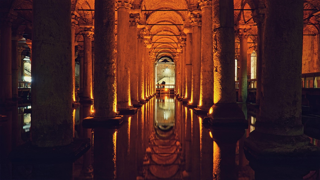Eine dramatisch beleuchtete Innenansicht der Basilica Cistern (Yerebatan Sarnıcı) in Istanbul, die perfekt das Thema "Mystik unter der Erde: Warum du die Basilica Cistern (Yerebatan Sarnıcı) neu entdecken musst" illustriert. Reihen massiver, alter Steinsäulen erstrecken sich in die Tiefe und spiegeln sich intensiv in der ruhigen Wasserfläche am Boden, wodurch eine geheimnisvolle, fast unendliche Perspektive entsteht, beleuchtet von warmen, bernsteinfarbenen Lichtern.