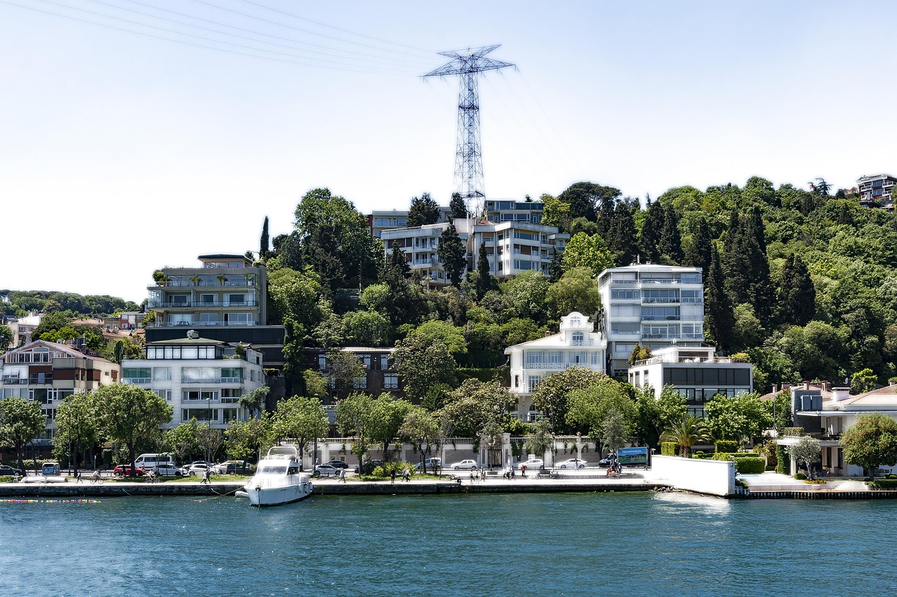 Blick auf die luxuriöse Küstenlinie am Bosporus mit eleganten Villen und modernen Apartmenthäusern, die sich einen dicht bewaldeten Hang hinaufziehen. Im Vordergrund ankert eine weiße Yacht im tiefblauen Wasser. Dieses Bild fängt die Atmosphäre eines Spaziergangs ein, wie er im Artikel "Bosporus-Flaneure: Mein Guide für einen Spaziergang von Arnavutköy nach Bebek" beschrieben wird, und zeigt die attraktive Architektur entlang dieser begehrten Strecke in Istanbul.