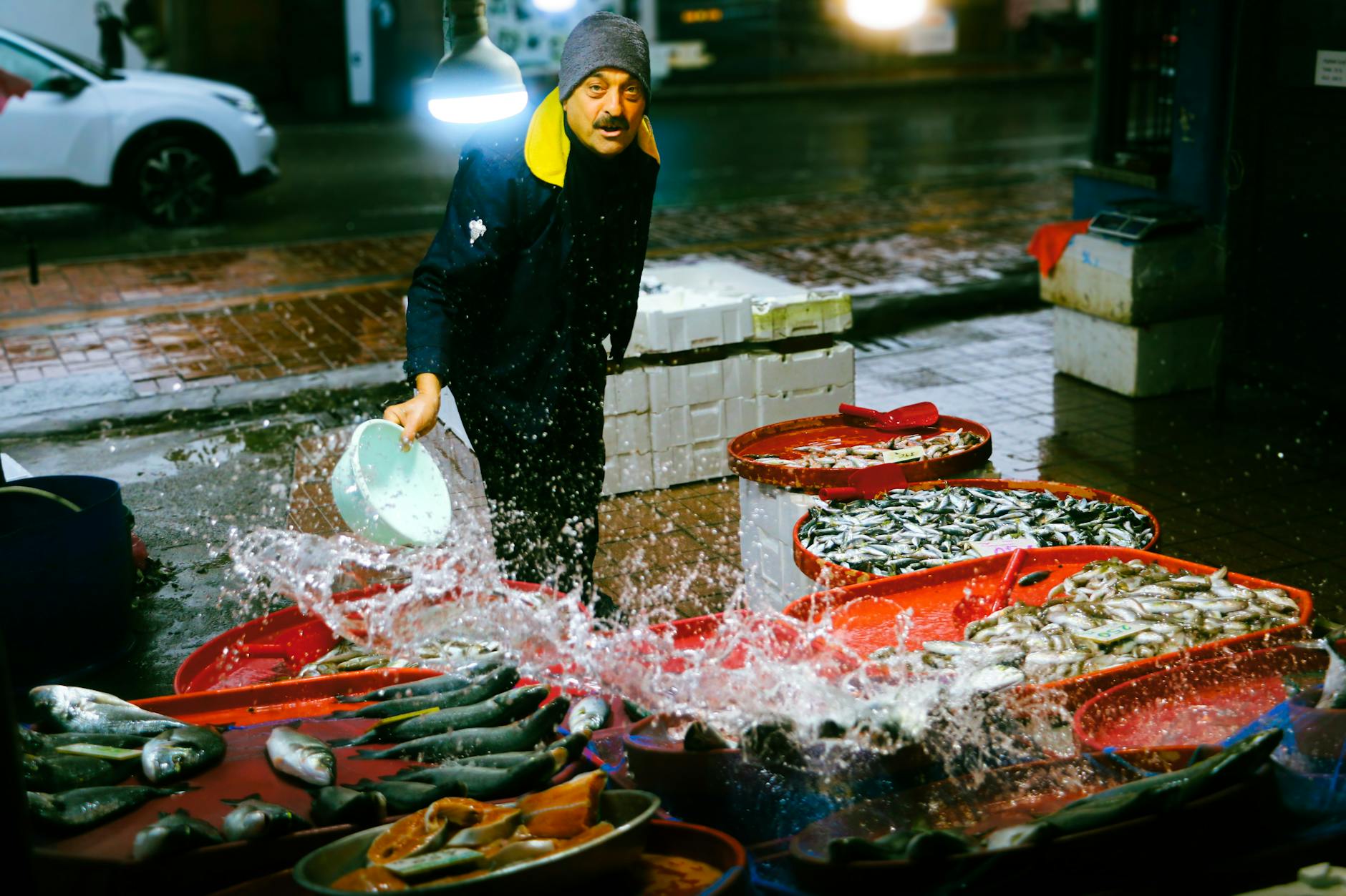 Ein lebendiges Nachtbild eines Fischverkäufers auf einem Istanbuler Markt, der mit einem Eimer Wasser über seinen frischen Fang spritzt, ein zentrales Element für jeden Guide zur 'Ehrliche Küche: Mein Guide zu den besten Esnaf Lokantası in Istanbul'. Mehrere rote Behälter sind mit glänzendem Fisch gefüllt, während im Hintergrund ein Mann mit Mütze und gelbem Kragen in die Kamera blickt, umgeben von nassen, beleuchteten Straßenoberflächen.