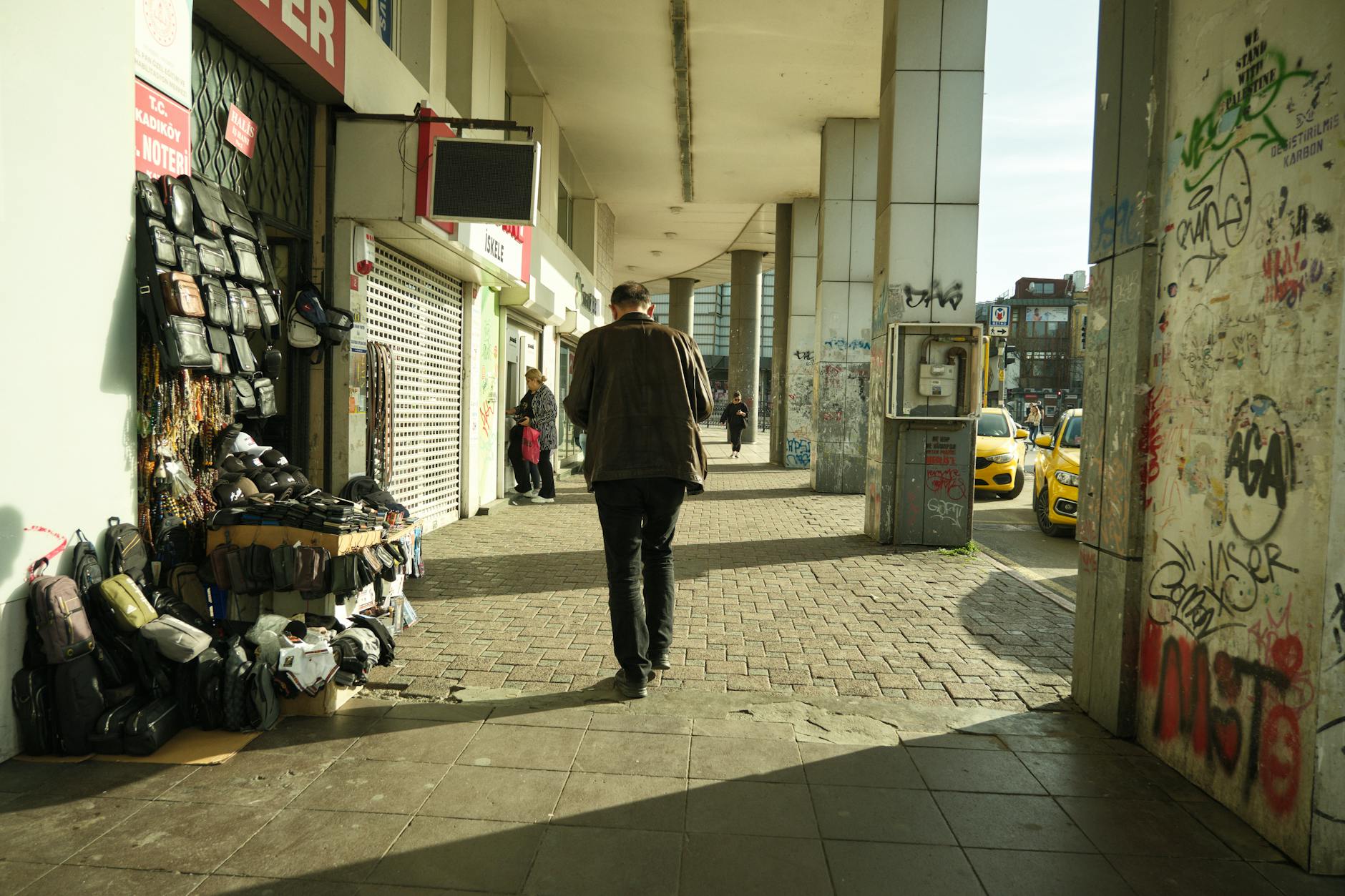 Blick auf einen Bürgersteig mit Ladenfronten und einem Straßenhändler in Kadıköy, Istanbul, mit stark bekritzelten Säulen.