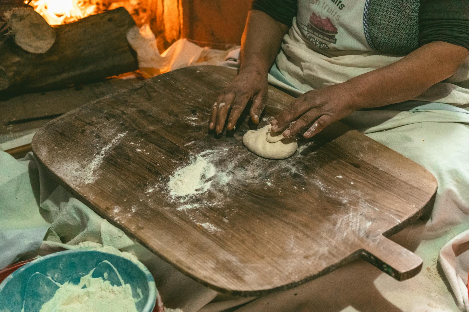 Eine Frau knetet traditionellen Teig auf einem Holzbrett vor einem wärmenden Kaminfeuer.
