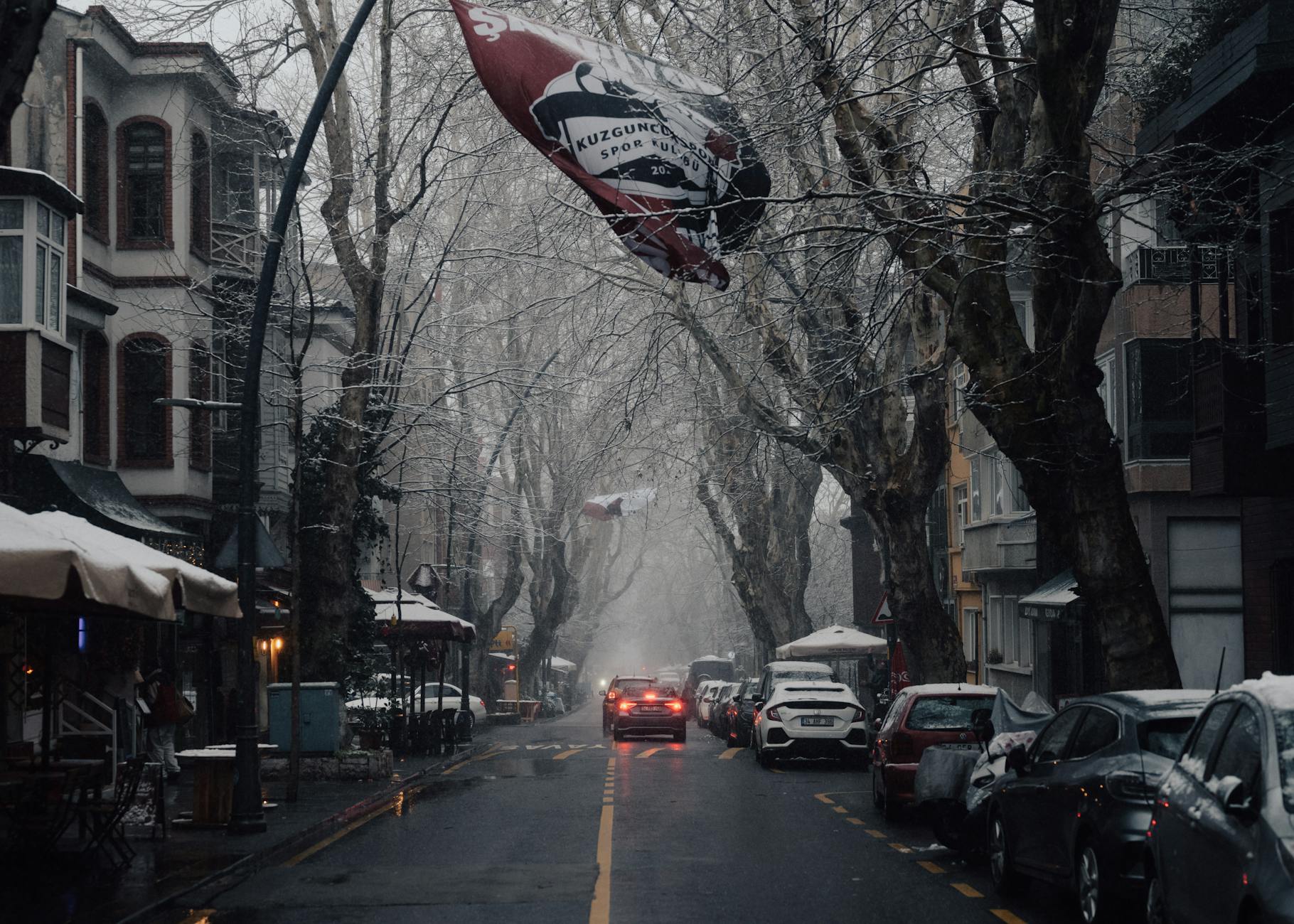 Schneebedeckte Straße in Kuzguncuk mit alten Bäumen und einer wehenden Fenerbahçe-Flagge mit der Aufschrift 'KUZGUNCUK SPOR KULÜBÜ'.